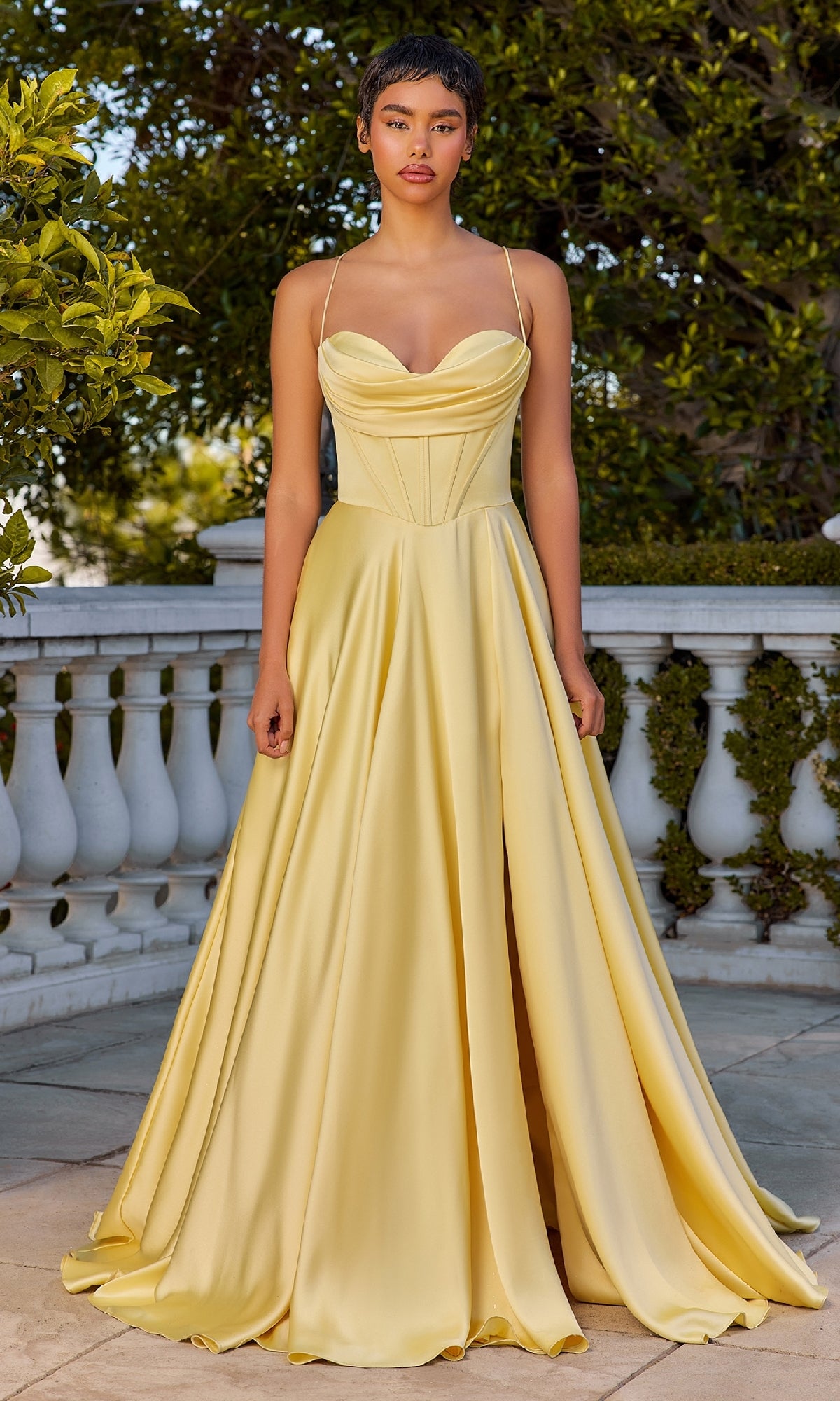 A woman stands outdoors in the Ladivine 7481 Long Satin A-Line Prom Dress, a floor-length yellow gown with thin straps and a fitted bodice, set against green trees and a white balustrade.